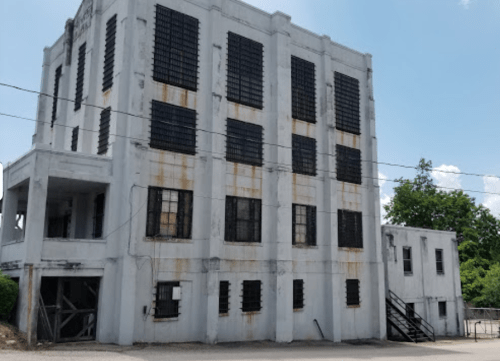 Abandoned white building with black window grates, showing signs of wear and rust, surrounded by greenery.