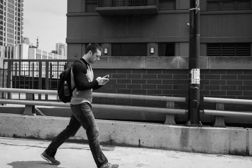 A man walks along a city sidewalk, focused on his phone, with a backpack slung over one shoulder.