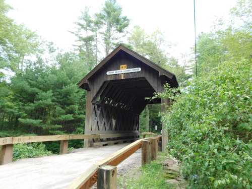 A wooden covered bridge with a sign, surrounded by trees and greenery, leading over a small path.