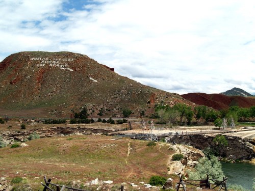 A scenic view of a mountain with "World's Largest Mineral Hot Springs" written on it, surrounded by greenery and a river.