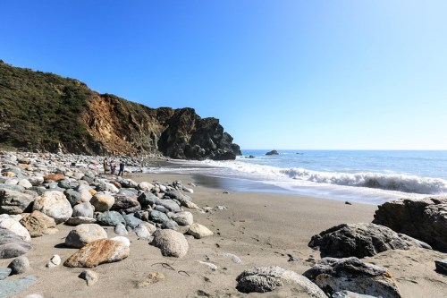 A rocky beach with gentle waves, surrounded by cliffs and clear blue skies, with a few people walking along the shore.