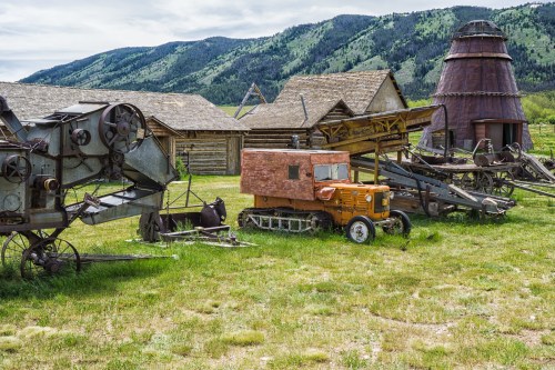 Rustic farm equipment and a vintage tractor are displayed in a grassy area, with mountains in the background.