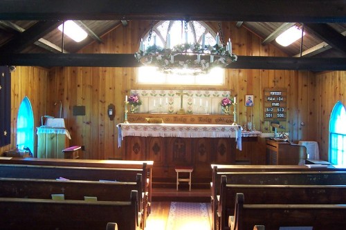 Interior of a rustic church with wooden pews, an altar adorned with flowers, and stained glass windows.