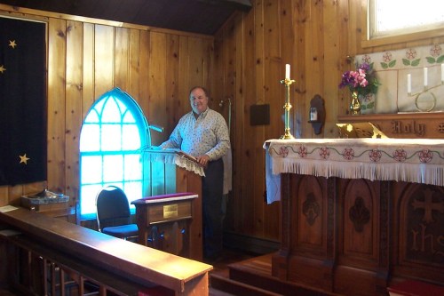 A man stands at a lectern in a wooden church interior, with an altar and stained glass window in the background.