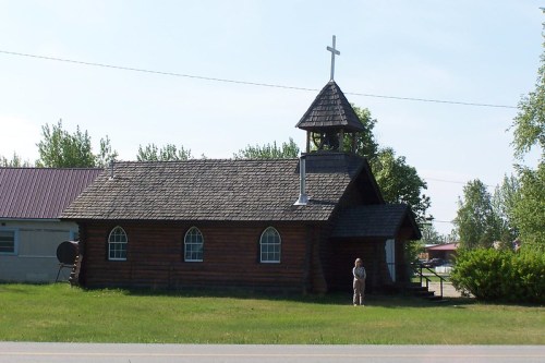 A small wooden church with a steeple and cross, surrounded by grass and trees, with a person standing nearby.