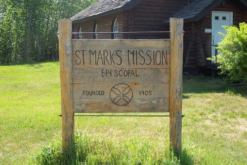 Sign for St. Mark's Mission, established in 1905, with a wooden structure and grassy surroundings.