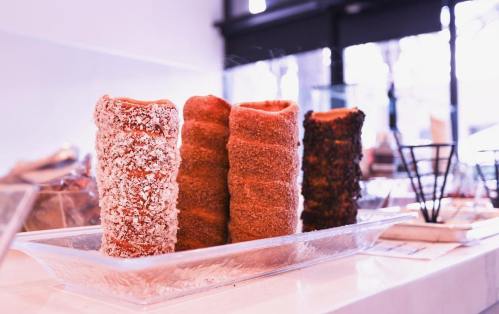 Three cylindrical pastries coated in different toppings, displayed on a glass plate in a bright café setting.