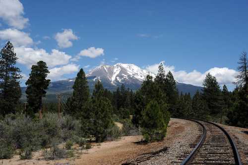 A scenic view of a snow-capped mountain with a railway track curving through a forested landscape under a blue sky.