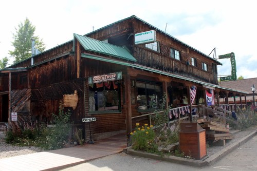A rustic wooden building with a green roof, featuring a gift shop and an open sign, surrounded by flowers and flags.