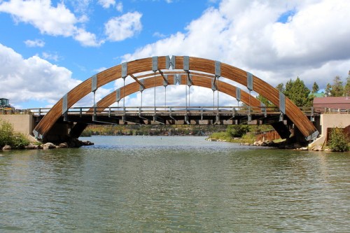 A wooden arch bridge spans a calm river, surrounded by trees and under a partly cloudy sky.