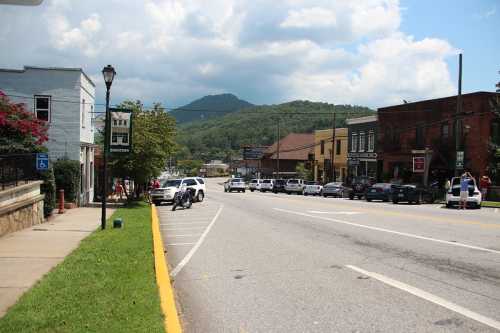 A small town street lined with shops, cars parked along the road, and mountains in the background under a cloudy sky.