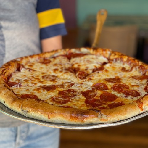 A person holds a large pepperoni pizza on a metal tray, showcasing its melted cheese and golden crust.