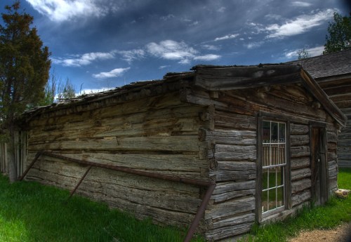 A rustic wooden cabin with a window, surrounded by green grass under a cloudy sky.