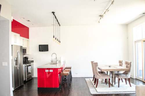 Modern kitchen and dining area featuring red accents, stainless steel appliances, and a wooden dining table with upholstered chairs.