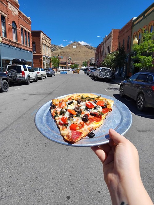 A hand holds a slice of pizza on a plate, with a sunny street and mountains in the background.