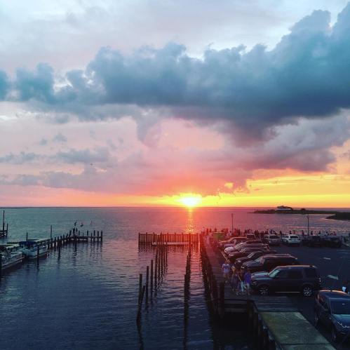 A vibrant sunset over a calm harbor, with boats docked and cars parked along the waterfront.