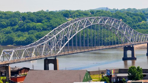 A large steel arch bridge spans a river, surrounded by green trees and a clear blue sky.