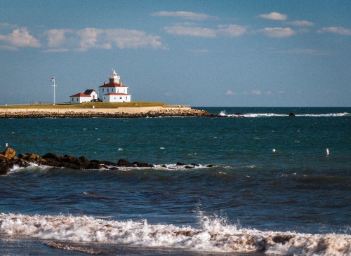 A lighthouse on a grassy shore, surrounded by blue water and gentle waves under a clear sky with scattered clouds.