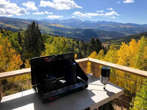 A camping stove on a table with a scenic mountain view and colorful autumn trees in the background.