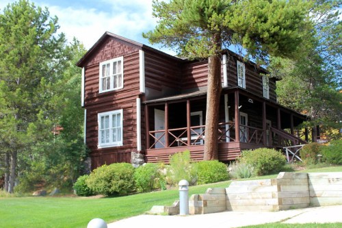 A rustic wooden house with a porch, surrounded by greenery and trees, set against a clear blue sky.