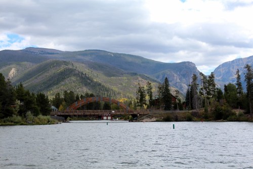 A serene lake surrounded by mountains, featuring a wooden bridge and lush trees under a cloudy sky.
