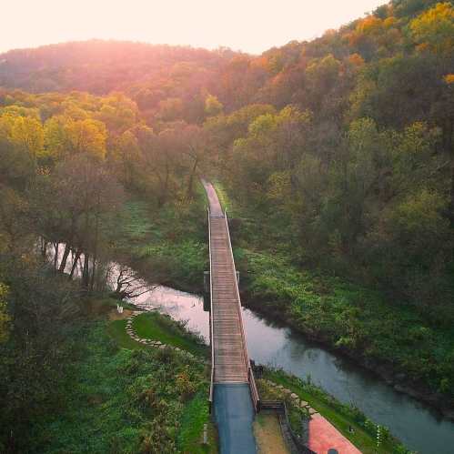 A scenic view of a wooden bridge over a river, surrounded by lush green trees and a sunset in the background.