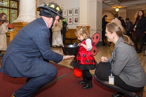 A man in a firefighter helmet kneels to give candy to a little girl in a ladybug costume, while a woman watches.