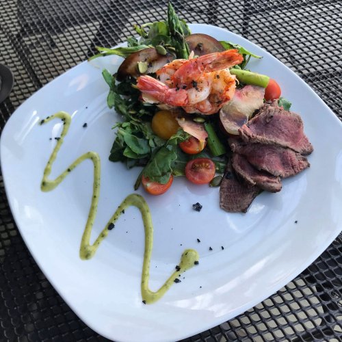 A vibrant salad with shrimp, cherry tomatoes, asparagus, and sliced beef, drizzled with green sauce on a white plate.