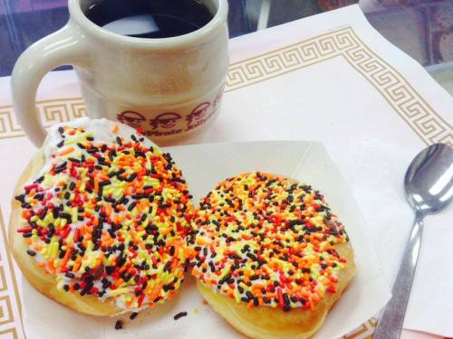 A cup of black coffee next to two colorful donuts topped with sprinkles on a decorative paper.