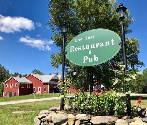 A green sign reading "the inn Restaurant & Pub" surrounded by flowers, with a red barn in the background under a blue sky.