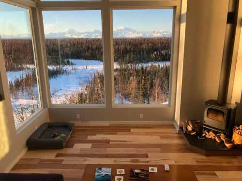 Cozy interior with large windows showcasing a snowy landscape and mountains in the distance, featuring a wood stove.