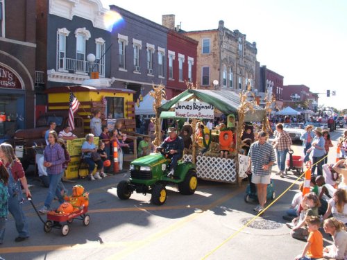 A festive parade scene with a decorated float, a tractor, and people enjoying the event on a sunny day.