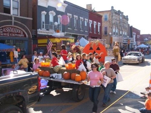 A festive parade scene with children on a float decorated with pumpkins and Halloween themes, surrounded by spectators.