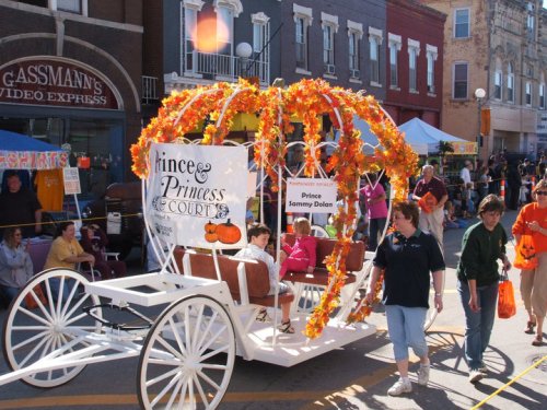 A decorated pumpkin carriage with children dressed as a prince and princess, parading at a festive event.