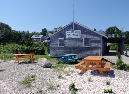 A rustic wooden building with a sign, surrounded by picnic tables and greenery on a sunny day.