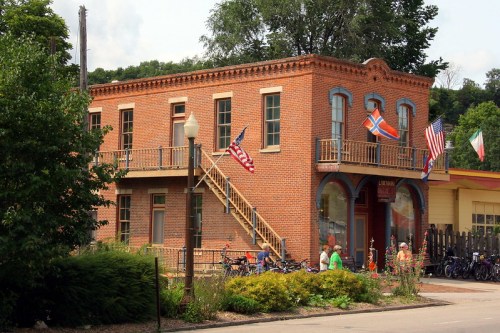 Historic brick building with flags, a staircase, and greenery in front, set in a quaint town.