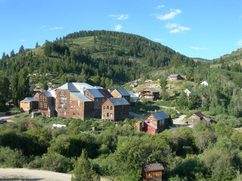 A scenic view of a rustic mountain town with wooden buildings surrounded by lush greenery and hills under a clear blue sky.