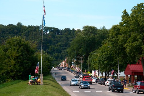 A scenic view of a small town street lined with trees, flags, and parked cars on a sunny day.