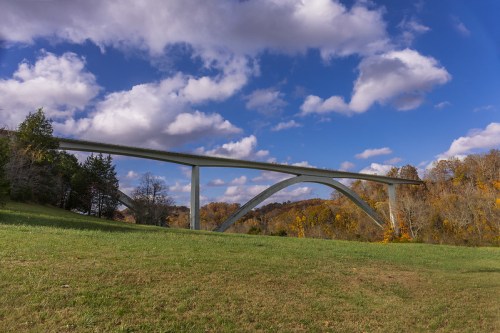 A modern bridge arches over a grassy field, surrounded by trees and a blue sky with fluffy clouds.