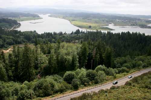 Aerial view of a winding river surrounded by lush greenery and a road, with distant hills under a cloudy sky.