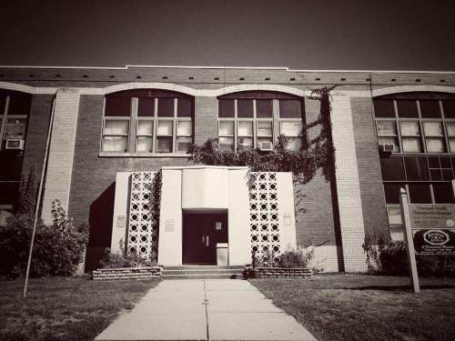 Black and white photo of a brick school building with large windows and a decorative entrance. Ivy grows on the facade.
