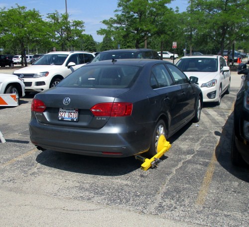 A gray Volkswagen sedan with a yellow wheel clamp parked in a lot, surrounded by other vehicles.