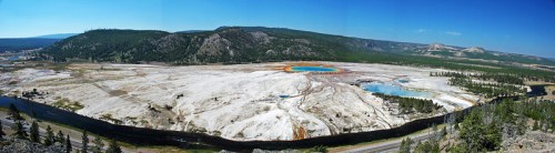 A panoramic view of Yellowstone National Park featuring colorful hot springs and surrounding green hills under a clear blue sky.