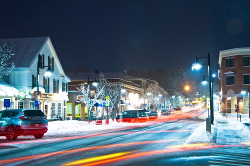 A snowy street at night, lined with shops and illuminated by streetlights, with car lights creating streaks.