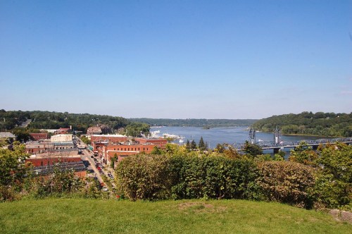 A scenic view of a river town with buildings, trees, and a bridge under a clear blue sky.