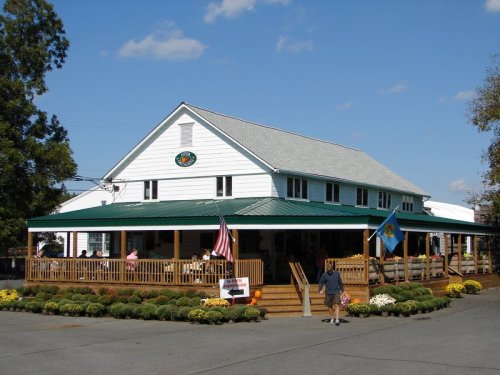 A large white building with a green roof, featuring a porch and flags, surrounded by flowers and trees on a sunny day.