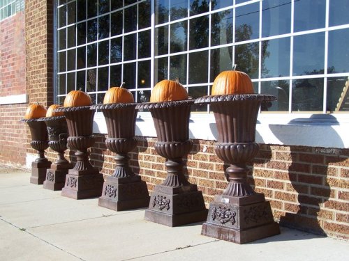 Five decorative urns lined up against a brick wall, each topped with a bright orange pumpkin.