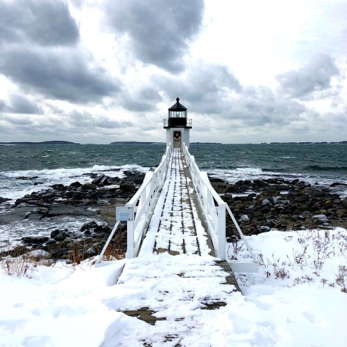 A snowy lighthouse stands on a rocky shore, with dark clouds and a calm sea in the background.