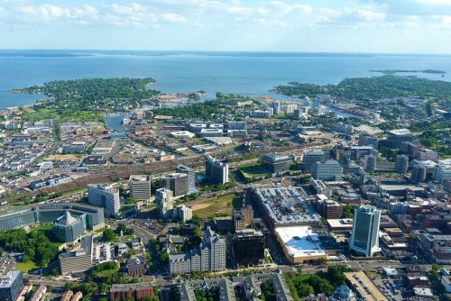 Aerial view of a city by the water, featuring buildings, roads, and green spaces with a distant island visible.