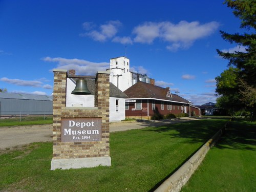 A brick sign for the Depot Museum, with a bell, set against a blue sky and green grass. The museum building is in the background.